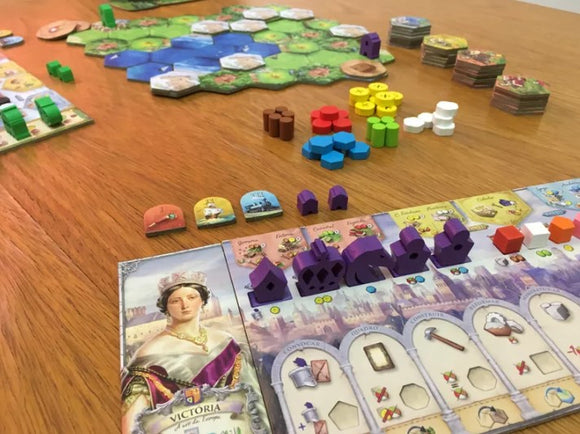 A vibrant board game setup for 'Brazil: Imperial' made by Portal. The game features colorful tokens in various shapes, including houses, resources, and action cards displayed on a wooden table. An illustrated character named Victoria is prominently featured on the game board, surrounded by green hexagonal tiles representing terrain and water areas, highlighting an engaging strategy game experience.
