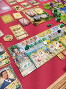 A vibrant board setup for the game 'Brazil: Imperial' by Portal. The image showcases various colorful game components including cards, tokens, and boards arranged on a red game table. Notable elements include characters and decorative tokens, highlighting the game's focus on strategy and resource management in Brazilian history. Players can engage with diverse mechanics as they navigate the game's rich theme.
