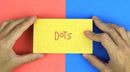 A person holding the FlipBoku: Dots product, which is a bright yellow box featuring embossed designs and the word 'Dots' in a playful red font. The background is split into two vibrant colors: red on the left and blue on the right.