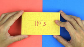 A person holding the FlipBoku: Dots product, which is a bright yellow box featuring embossed designs and the word 'Dots' in a playful red font. The background is split into two vibrant colors: red on the left and blue on the right.