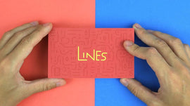 A person holding the 'Lines' product by Boku Flipbooks. The box has a textured red surface with the word 'LINES' in bold gold letters. The background features contrasting blue and red surfaces, enhancing the vibrant appearance of the product.