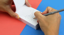 A person is holding a FlipBoku: Lines flipbook while sketching molecular structures in it with a pencil. The background features vibrant red and blue colors.
