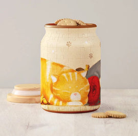 A vibrant 3D jigsaw puzzle designed as a jar, featuring a cozy scene of a sleeping orange cat surrounded by yarn and small yellow chicks. The jar has a lid resembling a cookie on top and is displayed on a light wooden surface with some decorative biscuit-shaped items nearby.