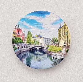 16-piece magnetic jigsaw puzzle featuring a scenic view of Ljubljana, Slovenia. The image showcases colorful buildings along a river with a clear blue sky above, inviting creativity and relaxation.