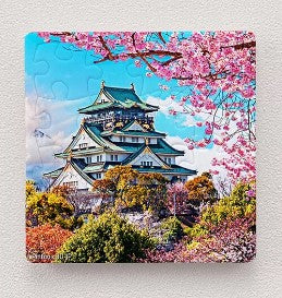 A vibrant 16-piece jigsaw puzzle featuring the iconic Osaka Castle surrounded by colorful cherry blossoms and lush greenery under a clear blue sky. The intricate design showcases the beauty of the castle in spring.