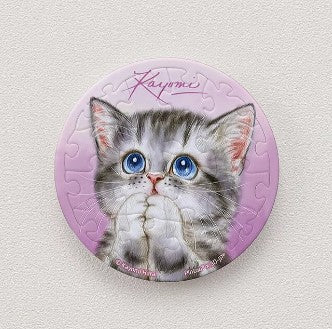 A round, jigsaw puzzle magnet featuring an adorable cat with blue eyes and a gray striped coat, set against a soft pink background. The design includes the name 'Kayomi' elegantly written at the top, conveying a sense of charm and whimsy. This 16-piece puzzle magnet can be easily displayed and is perfect for cat lovers.