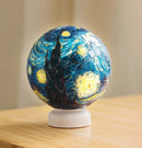 Close-up of the Van Gogh 'Starry Night' 3D jigsaw puzzle light featuring a spherical design with vibrant colors, including blue and yellow swirls, and a silhouette of a tower. The puzzle sits on a white base, showcasing the iconic artwork in a unique 3D form.