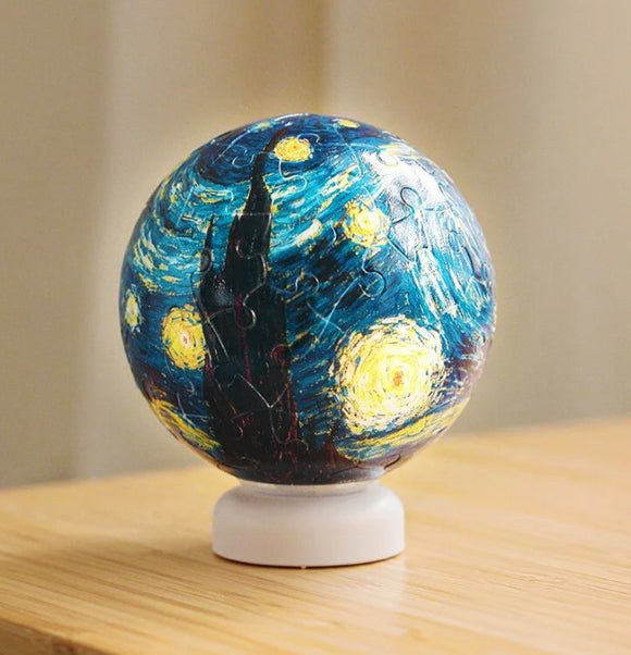 Close-up of the Van Gogh 'Starry Night' 3D jigsaw puzzle light featuring a spherical design with vibrant colors, including blue and yellow swirls, and a silhouette of a tower. The puzzle sits on a white base, showcasing the iconic artwork in a unique 3D form.