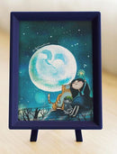 A completed jigsaw puzzle titled 'Starry Starry Night' featuring a whimsical scene with a girl and two cats watching a large full moon against a starry sky. The puzzle is displayed in a navy blue frame with a sturdy stand, perfect for home decor or gifting. Ideal for puzzle enthusiasts and art lovers alike.