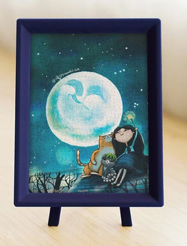 A completed jigsaw puzzle titled 'Starry Starry Night' featuring a whimsical scene with a girl and two cats watching a large full moon against a starry sky. The puzzle is displayed in a navy blue frame with a sturdy stand, perfect for home decor or gifting. Ideal for puzzle enthusiasts and art lovers alike.