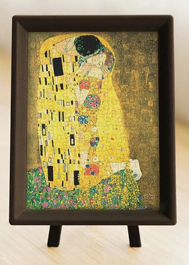 A completed 150-piece jigsaw puzzle of Gustav Klimt's famous painting 'The Kiss'. The puzzle is displayed in a framed stand, showcasing vibrant colors and intricate designs, highlighting the intimate embrace of the couple surrounded by golden hues and floral patterns.