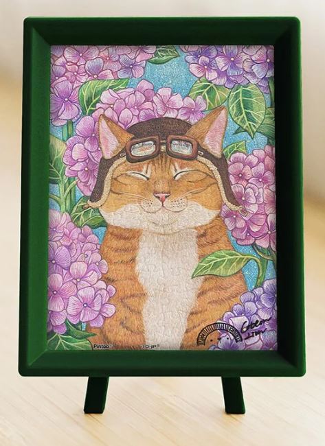A vibrant jigsaw puzzle featuring a cheerful cat wearing aviator goggles, surrounded by beautiful purple hydrangea flowers. The puzzle is displayed in a green frame with a stand, showcasing the playful artwork of a whimsical feline character, making it a charming addition to any puzzle collection.