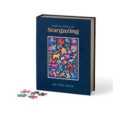 The Constellations 101 Book Puzzle by Galison features a vibrant illustration of various constellations against a dark blue background. The puzzle is in a book-like format, with a colorful, eye-catching image on the cover and scattered jigsaw pieces nearby, highlighting its 500-piece count. Perfect for stargazing enthusiasts and puzzle lovers alike.