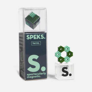The Speks Elements: Terra is displayed in its transparent packaging, featuring a small magnetic puzzle made up of green and teal beads. The product showcases an artistic arrangement of magnet balls forming a geometric shape next to the box, emphasizing its creative potential and tactile interaction. The packaging prominently features the SPEKS logo and the words 'spectacularly magnetic' on the side.