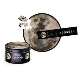 The Tin Space Puzzle Moon features a round jigsaw puzzle depicting a detailed image of the moon, with some pieces around it and a faux photo tin container. It includes a guide on moon phases, showcasing different lunar appearances.
