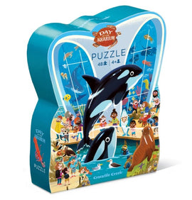 The 'Day at the Aquarium' jigsaw puzzle by Crocodile Creek features a colorful scene depicting various sea animals, including orcas and a walrus, with children and adults enjoying their time at the aquarium. The puzzle comes in a uniquely shaped box, making it an engaging and cute addition for kids aged 4 and up.