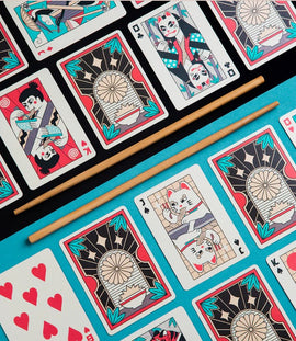 A vibrant set of Ramen Heads playing cards featuring colorful illustrations. The cards showcase whimsical designs inspired by ramen culture, including cats and characters enjoying noodles. A pair of wooden chopsticks is placed centrally on a bright turquoise background, enhancing the playful theme of the game.