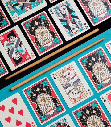 A vibrant set of Ramen Heads playing cards featuring colorful illustrations. The cards showcase whimsical designs inspired by ramen culture, including cats and characters enjoying noodles. A pair of wooden chopsticks is placed centrally on a bright turquoise background, enhancing the playful theme of the game.
