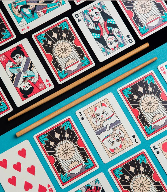 A vibrant set of Ramen Heads playing cards featuring colorful illustrations. The cards showcase whimsical designs inspired by ramen culture, including cats and characters enjoying noodles. A pair of wooden chopsticks is placed centrally on a bright turquoise background, enhancing the playful theme of the game.