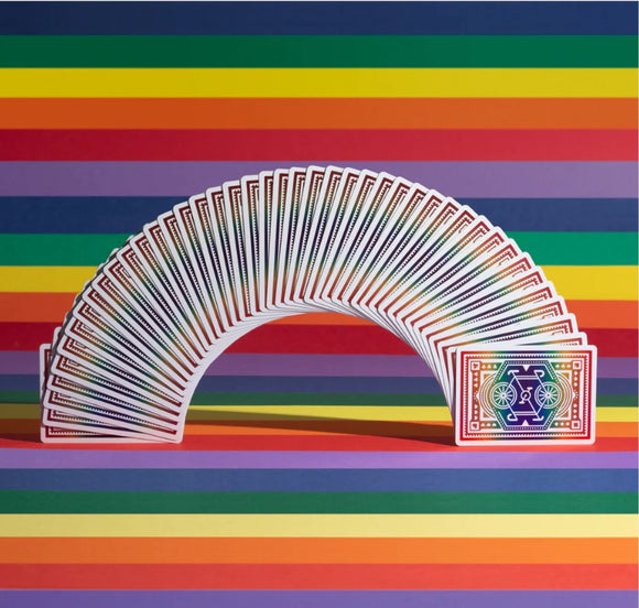 A vibrant display of DKNG Rainbow Wheels Playing Cards arranged in a fan shape, showcasing colorful card backs against a multi-colored striped background. The cards feature intricate designs, perfect for card games and collectors alike.