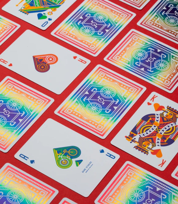 A vibrant display of DKNG Rainbow Wheels Playing Cards laid out on a red background. The deck features colorful designs of bicycles and unique illustrations on the face cards, showcasing a lively and artistic aesthetic ideal for card games.
