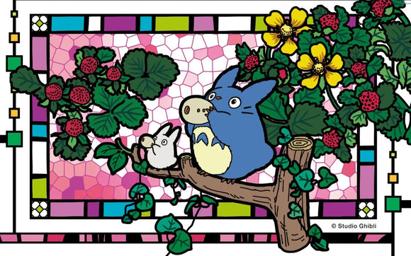 A whimsical jigsaw puzzle featuring Totoro playing the ocarina, surrounded by foliage and flowers. The colorful design includes details like strawberries and a stained glass frame, ideal for fans of Studio Ghibli.