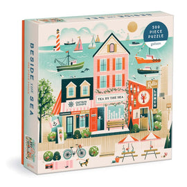 Galison's 'Beside the Sea' jigsaw puzzle features a charming coastal scene with colorful buildings, a vibrant beach atmosphere, and boats sailing in the water. The puzzle contains 500 pieces and is designed to provide an engaging and enjoyable challenge for puzzle enthusiasts.