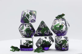 A Green Poison Frog RPG Dice Set by Foam Brain featuring seven unique polyhedral dice. Each die is transparent with vibrant green and purple colors, incorporating small green frog figures inside. The numbers are clearly visible, making them perfect for any role-playing game.