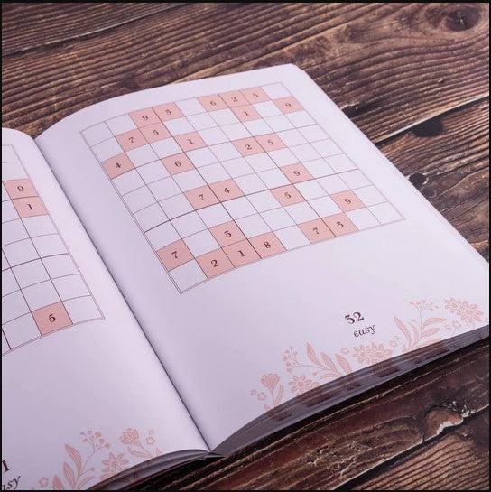 Open book page displaying a Sudoku puzzle. The grid shows several numbers filled in, along with empty boxes outlined in pink. The bottom corner indicates the puzzle is rated as "easy." A floral design is subtly featured along the edges of the page, adding a vintage touch to the overall aesthetic.