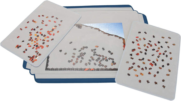 A close-up image of the Puzzle Pad featuring a 1500-piece jigsaw puzzle by Heye. Several puzzle pieces are scattered on two trays, while a partially completed section is visible on the main pad. The trays and pad are designed for organizing puzzle pieces, showcasing a variety of colors and shapes.