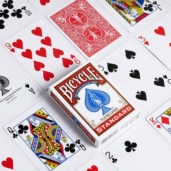 A standard deck of Bicycle playing cards featuring the iconic blue spade logo on the front, surrounded by various playing cards including hearts, diamonds, and spades laid out on a flat surface.