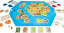 Catan: Seafarers board game featuring a colorful hexagonal game board with various terrains, player pieces in blue, red, yellow, and orange, resource cards, dice, and development cards arranged around the board, all set on a white background.