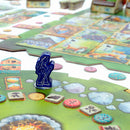 A colorful board game titled 'Meadow' by Rebel, featuring a blue game piece on a vibrant green landscape board. The game board is adorned with various tiles depicting animals and nature, alongside different tokens used for gameplay. This engaging game emphasizes strategy and exploration.