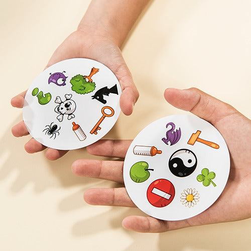 Two hands holding circular cards from the Spot it! game by Zygomatic. Each card features a variety of colorful images including a spider, a key, a green creature, and a yin-yang symbol. The game encourages players to quickly spot matching symbols between cards, promoting fast visual recognition and reaction skills. Perfect for family game nights, parties, or casual fun.