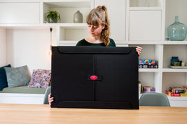 A young woman holds a large black storage case for jigsaw puzzles, labeled with a red emblem. The background shows a bright, modern living space with shelves containing various games and colorful items.