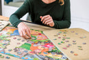 A person assembling the PortaPuzzle Standard 1500pc jigsaw puzzle by Jumbo, featuring vibrant artwork. The puzzle pieces are spread out on a table, showcasing a colorful scene with various characters and elements. The individual's hand is reaching for a puzzle piece, demonstrating an engaging and leisurely activity.