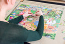 A person carefully assembling the PortaPuzzle Standard 1500pc jigsaw puzzle produced by Jumbo. The colorful puzzle features a lively farm scene with various characters and detailed illustrations. The individual is focused, adding pieces to create the picture on a flat surface, surrounded by extra puzzle pieces. The setting is bright and comfortable, ideal for enjoying this relaxing activity.