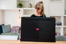 A woman holding the PortaPuzzle Standard, a black jigsaw puzzle storage board designed for 1500 pieces, showcasing its sleek design and functional features in a cozy room setting.