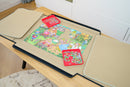 PortaPuzzle Standard 1500pc jigsaw puzzle laid out on a wooden table, featuring colorful artwork of whimsical characters and scenes. Two trays with scattered puzzle pieces are on either side, complemented by storage flaps to keep the puzzle organized.