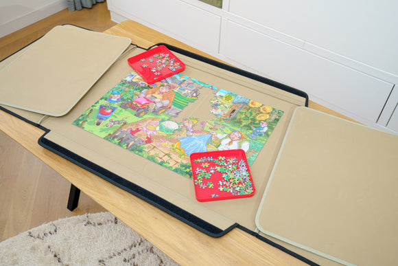 PortaPuzzle Standard 1500pc jigsaw puzzle laid out on a wooden table, featuring colorful artwork of whimsical characters and scenes. Two trays with scattered puzzle pieces are on either side, complemented by storage flaps to keep the puzzle organized.