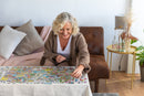 A woman with curly blonde hair smiles as she places a piece into the colorful Jigsaw puzzle titled 'Cattle Market' by Jan van Haasteren. The puzzle features a lively farm scene filled with animals and people, showcasing intricate details. She sits at a table in a cozy living space with a brown leather couch and green plants in the background.