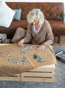 An older woman enjoying a jigsaw puzzle on a burlap-covered table. She is seated comfortably on the floor, with puzzle pieces scattered around her, focusing on assembling pieces. In the background, there are boxes of jigsaw puzzles stacked on a sofa.