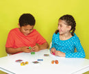 Two children, a boy wearing an orange shirt and a girl in a blue polka dot top, are having fun playing with colorful cube-shaped game pieces and cards on a white table. The background is bright yellow, creating a cheerful atmosphere.