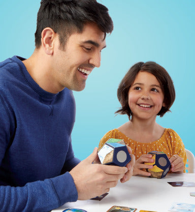 A father and daughter are playing the game 'Planet' by Blue Orange U.S.A. The father is smiling while holding a colorful geometric puzzle piece, and the daughter, wearing a yellow dress, is excitedly holding another piece. They are sitting at a table with cards and game pieces, with a light blue background enhancing the joyful atmosphere of family bonding.