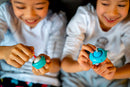 Two children smile as they play with the Big Bubble Cat Keychain, a colorful, playful fidget toy. One child holds a turquoise keychain while the other holds a matching light blue one. Both appear engaged and happy as they twist and interact with the toys, showcasing the fun and creativity in play.