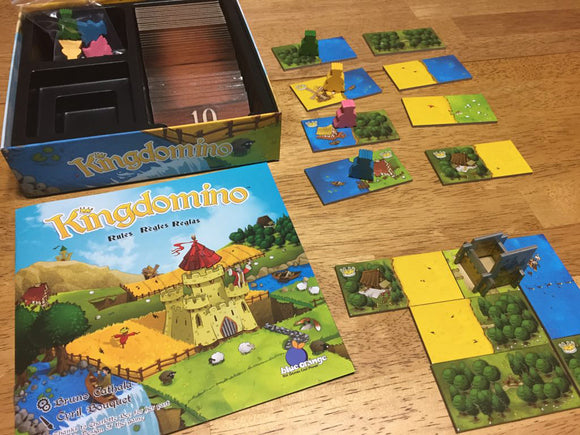 The Kingdomino board game by Blue Orange U.S.A. features a vibrant box cover depicting a whimsical kingdom with a castle, fields, and water. The game components include colorful domino tiles laid out on a wooden surface, with various landscapes and buildings illustrated on them. Pawns in different colors are placed on the tiles, enhancing the strategic gameplay experience.