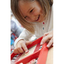 A young girl with light brown hair smiles while playing a game. She is focused on a colorful red game board that features a checker pattern, using her finger to interact with a game piece. The background is slightly blurred, showcasing a vibrant play area.