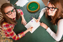 A Circle Wooden Dice Tray from Brybelly, designed to enhance game nights, is seen on a green felt-lined tabletop surrounded by colorful dice. Two young women, one in a plaid shirt and the other in a white sweater, are writing with pencils on paper, enjoying a fun gaming session together. The setup suggests a lively atmosphere perfect for family and friends.