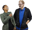 Two people, a man and a woman, are laughing and playfully interacting while wearing exaggerated fake moustaches. The woman, wearing a green jacket, appears joyful, while the man, in a blue shirt and black hoodie, smiles cheekily. Their playful expressions convey a light-hearted atmosphere.
