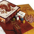 Tsuro board game by Calliope, featuring a beautifully designed box with a red dragon motif. The game includes colorful playing pieces and cards on a decorative game board. The rules are visible on one of the cards, surrounded by intricate designs.